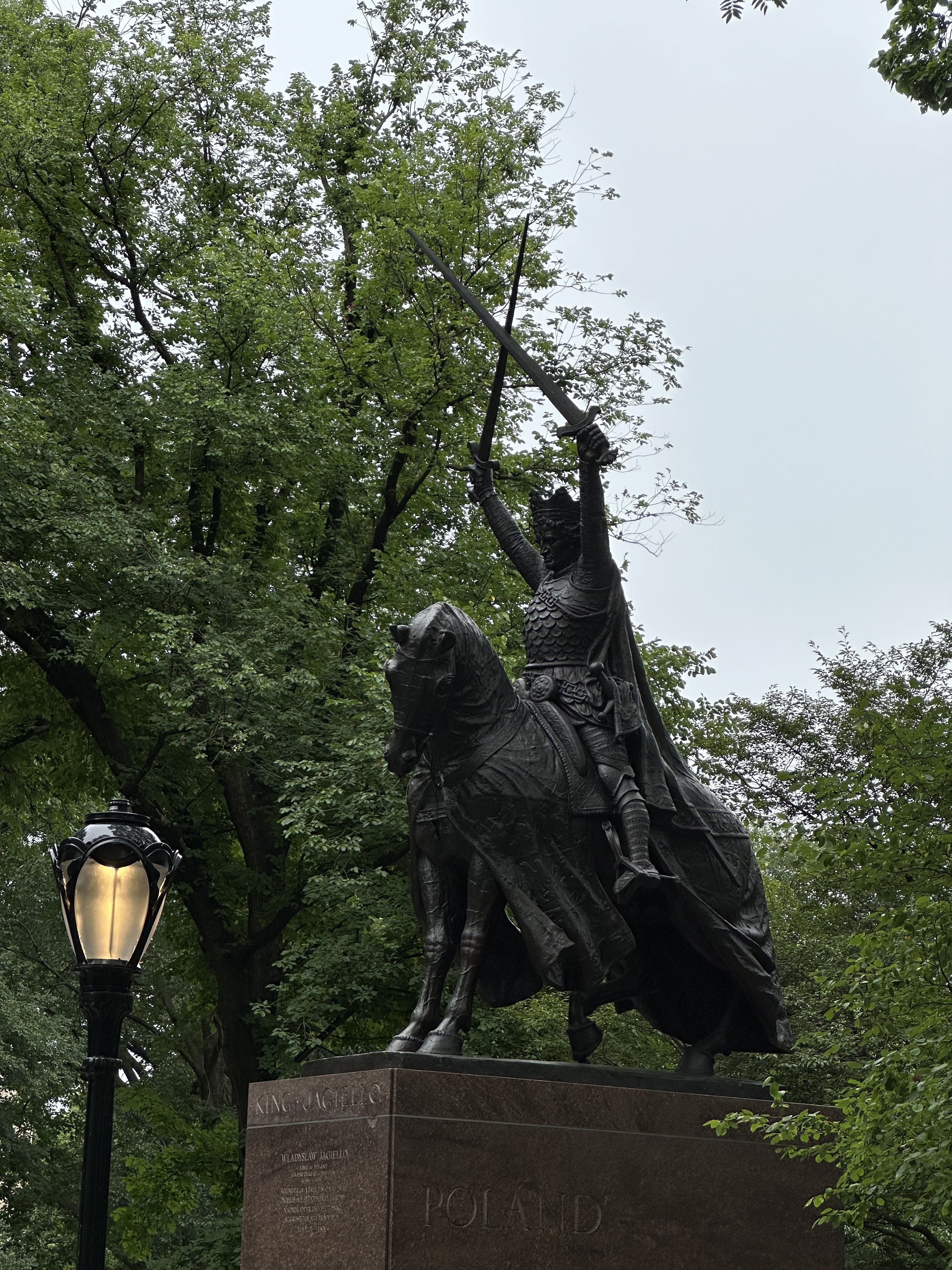 Equestrian statue of King Władysław II Jagiełło in Central Park, New York. The bronze statue depicts the king on horseback, raising two swords. The pedestal is inscribed with 'POLAND'. Trees and a lamppost are visible in the background.