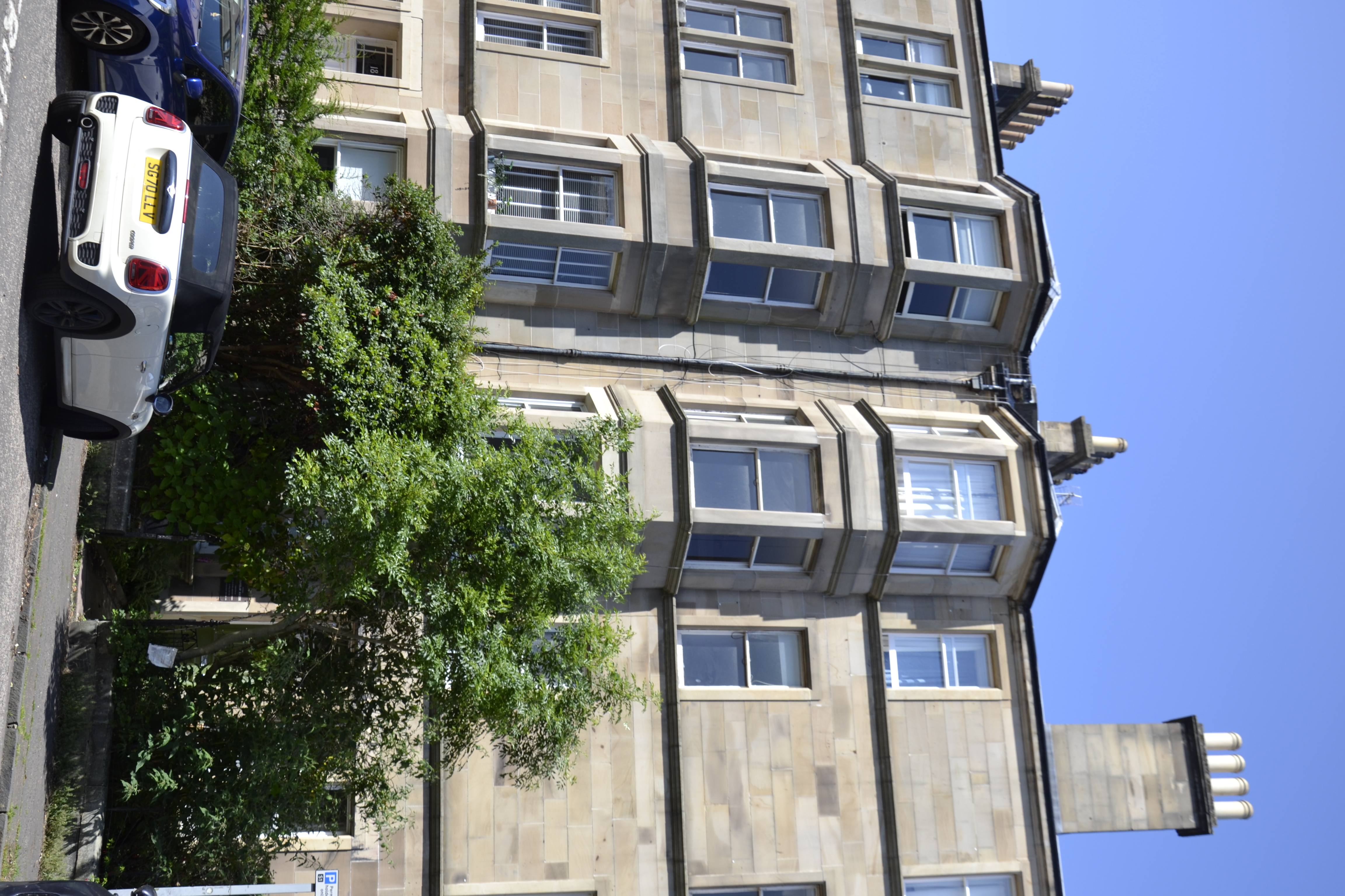 Un immeuble en grès du XIXe siècle dans le quartier de Marchmont à Édimbourg, avec des fenêtres en baie et une voiture décapotable blanche garée devant. Une végétation luxuriante cache partiellement l'entrée.
