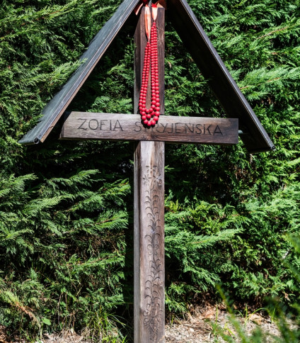 Drewniany krzyż na grobie Zofii Stryjeńskiej na cmentarzu w Chêne-Bourg, Genewa. Krzyż w stylu zakopiańskim z jej imieniem, ozdobiony czerwonym naszyjnikiem z koralików. Grób otacza zielona roślinność.