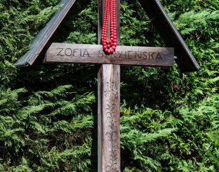 Drewniany krzyż na grobie Zofii Stryjeńskiej na cmentarzu w Chêne-Bourg, Genewa. Krzyż w stylu zakopiańskim z jej imieniem, ozdobiony czerwonym naszyjnikiem z koralików. Grób otacza zielona roślinność.