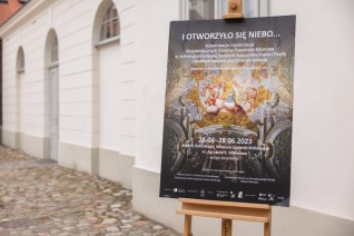 Plakat wystawy 'I otworzyło się niebo...' dotyczącej konserwacji fresków Franciszka Ecksteina we Lwowie, umieszczony na sztaludze przed budynkiem.