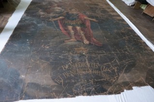 Obraz 'Święty Benedykt' na płaskiej powierzchni, widoczne ślady zużycia. Święty przedstawiony całopostaciowo z łacińskim napisem poniżej. Dzieło poddawane konserwacji.