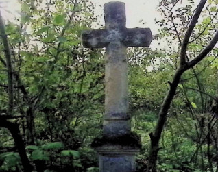 Kamienny krzyż na cokole otoczony gęstą zielenią. Krzyż ma ścięte krawędzie i spłaszczoną formę ostrosłupa na końcach ramion.