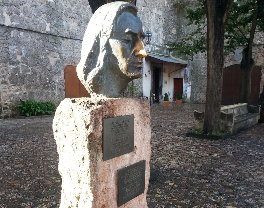 Popiersie Fryderyka Chopina z brązu na kamiennym cokole z tablicami, znajdujące się na dziedzińcu dawnego klasztoru kartuzów w Valldemossie na Majorce. W tle drzewa i kamienny mur.