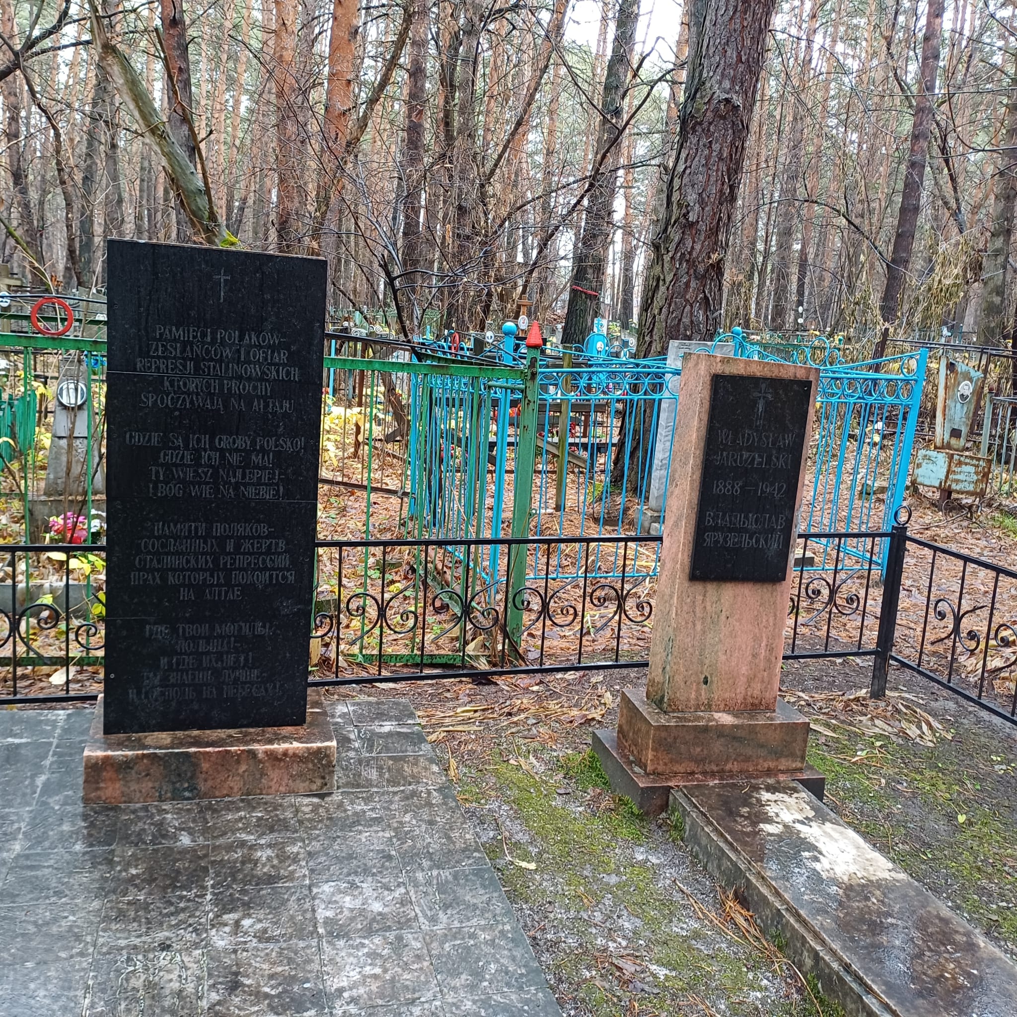 Two commemorative plaques in a forest cemetery, one dedicated to Polish exiles and victims of Stalinist repression, the other to Wladyslaw Jaruzelski. In the background a blue metal fence and trees.