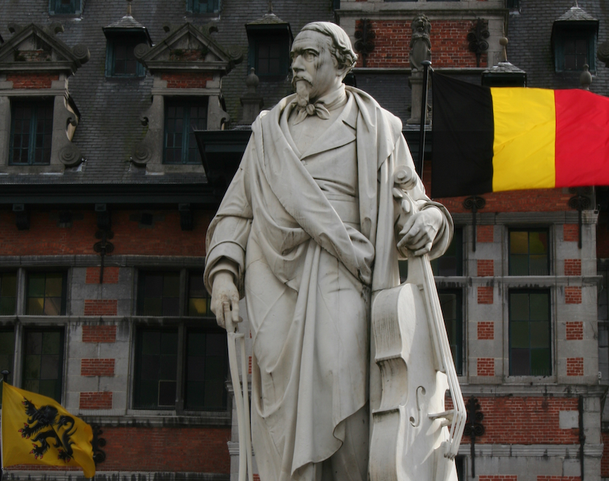 Statue of Adrien-François Servais holding a cello, on a pedestal with a laurel wreath, in front of a historic building with Belgian and Flemish flags in Halle.