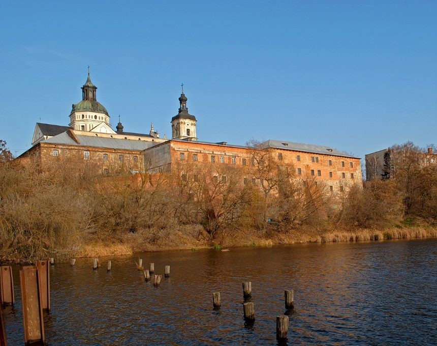 The Monastery of the Discalced Carmelites in Berdychiv, Ukraine, with its brick walls and towers, is seen from across the river. The foreground features water and wooden posts.