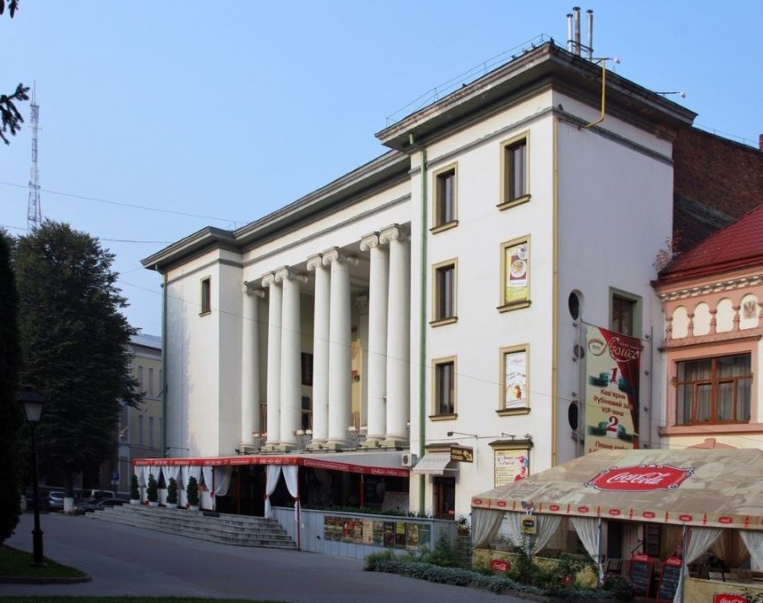 Bâtiment historique à Stanisławów avec une façade ornée de grandes colonnes et un café au rez-de-chaussée. La structure est adjacente à un bâtiment au toit rouge.