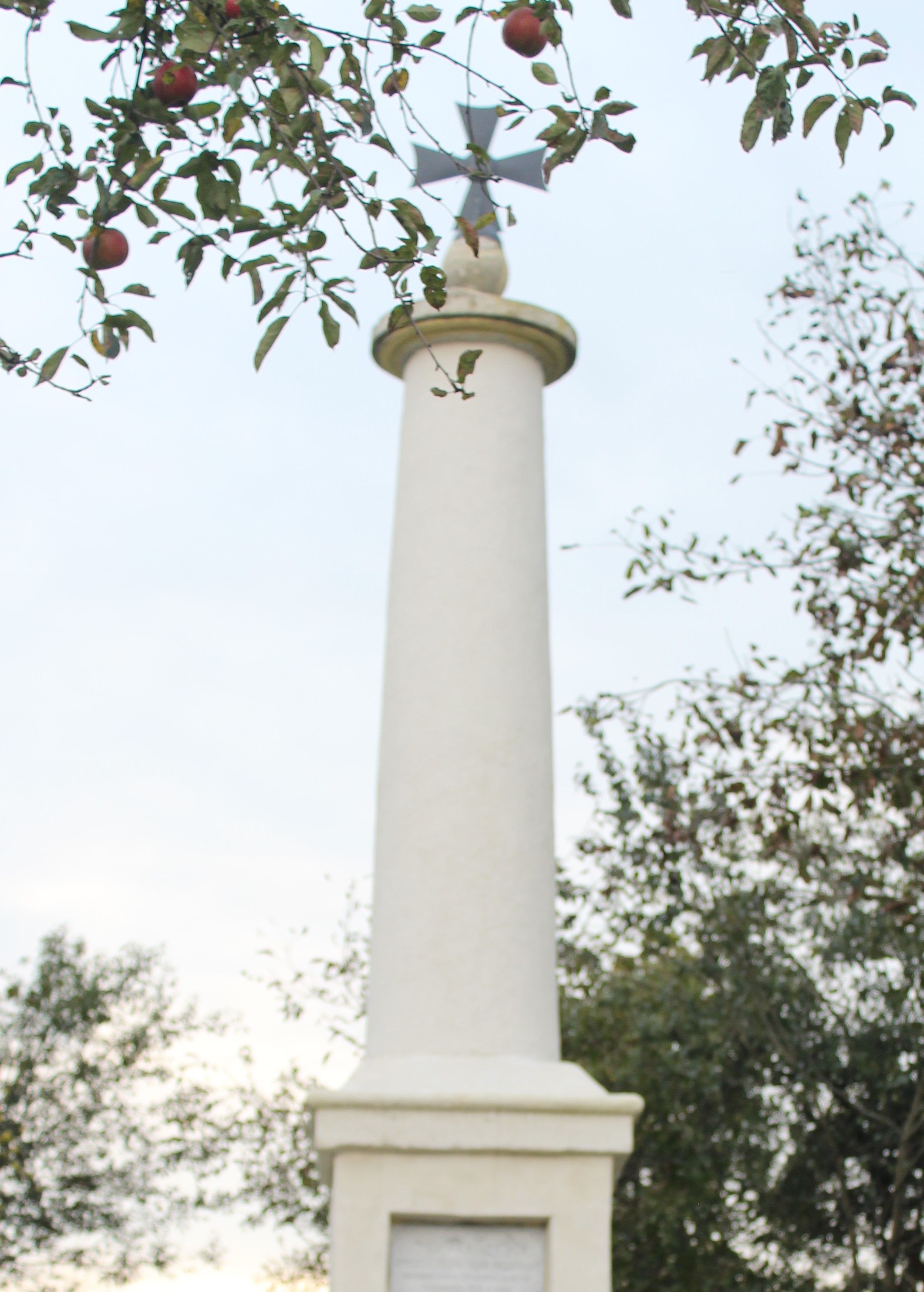 A tall white column with a cross on top, surrounded by trees with red apples.