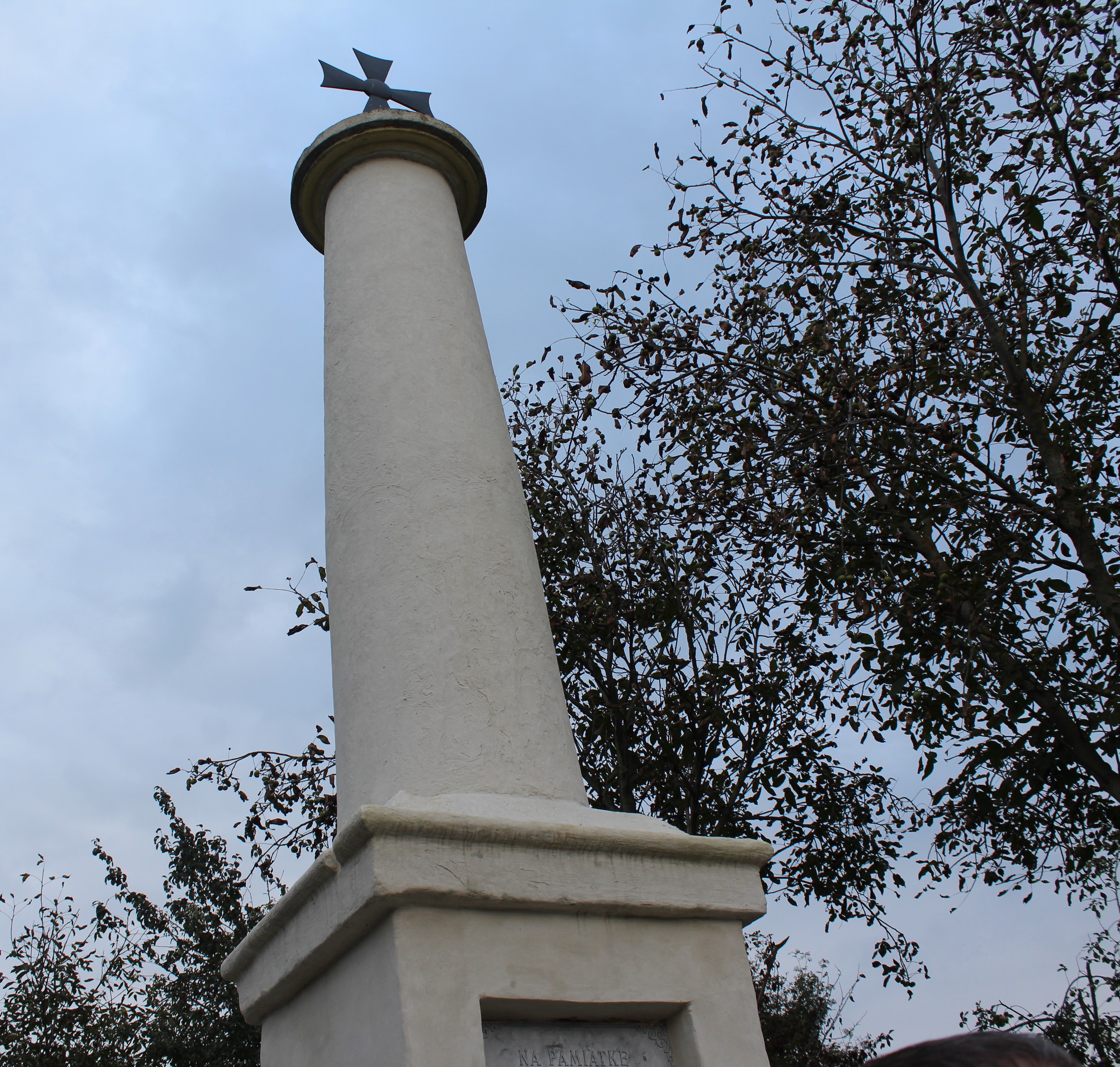 A tall, white column with a cross on top, surrounded by trees against a cloudy sky.