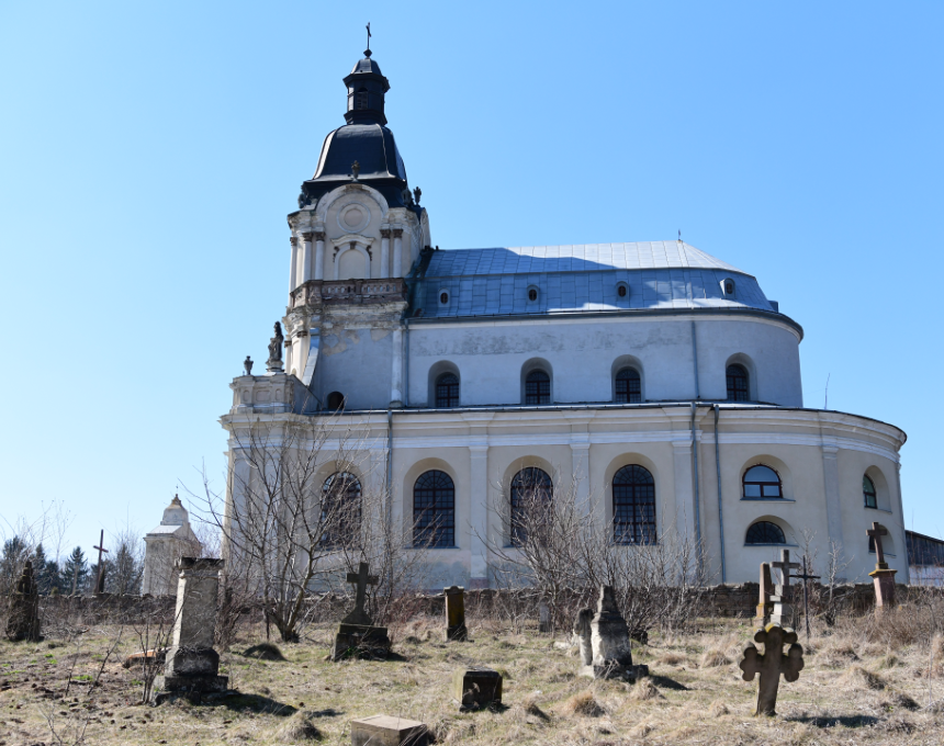 Widok boczny Kościoła Trójcy Świętej i klasztoru Misjonarzy w Mikulińcach, ukazujący barokową architekturę z kopułą i łukowatymi oknami. Na pierwszym planie widoczne stare nagrobki i krzyże.