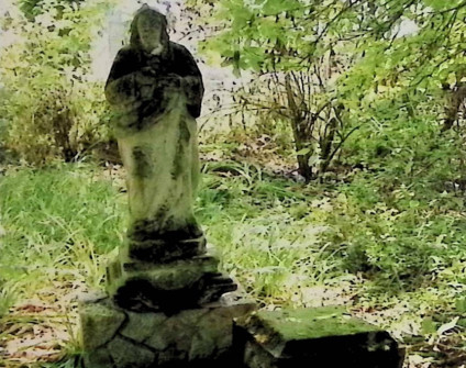 Zniszczona figura Chrystusa na dwustopniowym cokole w trawiastym, zalesionym terenie. Postać bosa, w przepasanej szacie i płaszczu, z dłońmi na piersi. Cokół przypomina skały.