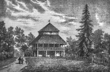 Stary lamus w Bali Wielkiej z drewnianą konstrukcją na kamiennych fundamentach, otoczony drzewami i ogrodem. Dwie osoby idą ścieżką prowadzącą do budynku.