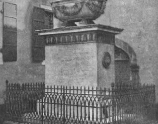 Pomnik generała Paca w Smirnie, z białym marmurowym sarkofagiem na cokole, otoczony żelaznym ogrodzeniem. Pomnik znajduje się na dziedzińcu z budynkiem w tle.