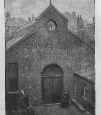 Kościół św. Józefa i św. Kazimierza w Londynie, budynek z cegły z krzyżem na szczycie, otoczony innymi budowlami. Osoba w ciemnym ubraniu stoi przy wejściu.