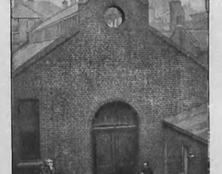 Kościół św. Józefa i św. Kazimierza w Londynie, budynek z cegły z krzyżem na szczycie, otoczony innymi budowlami. Osoba w ciemnym ubraniu stoi przy wejściu.