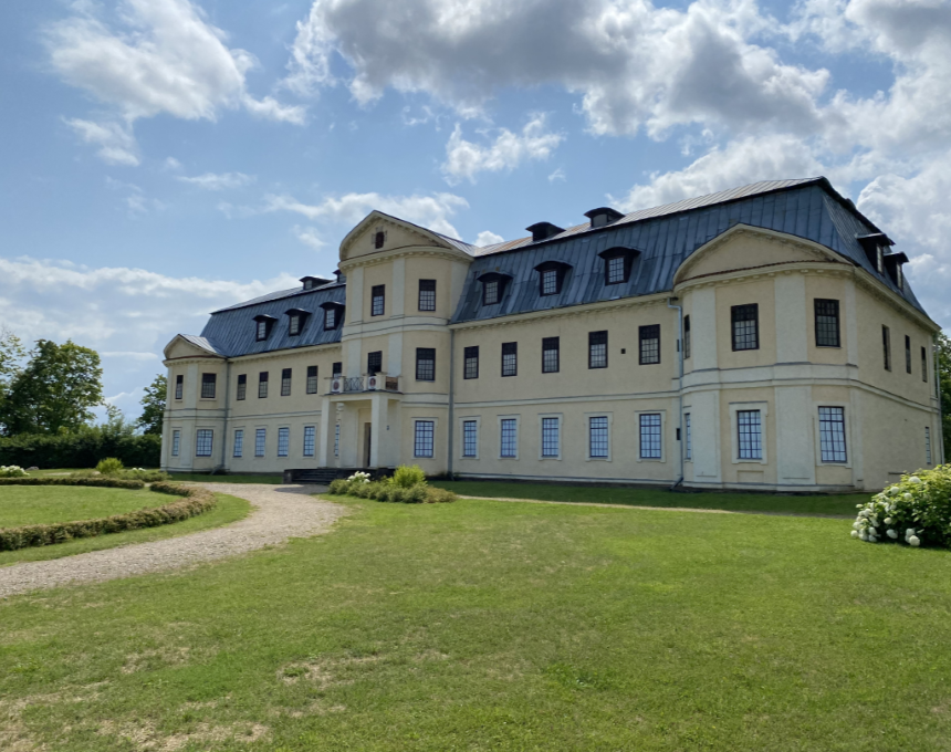 The Plater Palace in Kraslaw, a large, bright building with a mansard roof and numerous windows, surrounded by a green lawn and a path leading to the entrance.