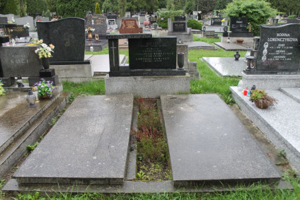 Miejsce pochówku z dwiema płaskimi płytami nagrobnymi i niską stelą na cokole z rytym krzyżem i inskrypcją. Widoczne otaczające groby i zieleń.