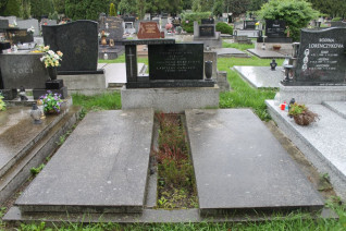 Miejsce pochówku z dwiema płaskimi płytami nagrobnymi i niską stelą na cokole z rytym krzyżem i inskrypcją. Widoczne otaczające groby i zieleń.