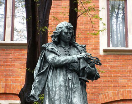 Brązowy posąg postaci historycznej w szacie, trzymającej sferę niebieską, na tle ceglanego budynku z oknami.