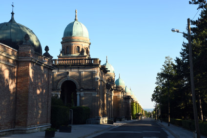 Cmentarz Mirogoj w Zagrzebiu, z widocznymi ceglanymi arkadami z zielonymi kopułami i alejką wśród drzew pod błękitnym niebem.