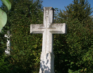 Kamienny krzyż na cokole z fazowanymi ramionami i mniejszym krzyżem na przecięciu. Krzyż stoi na tle gęstej zieleni.