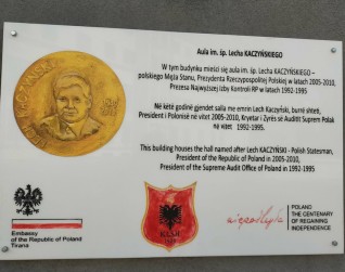 Trzy osoby odsłaniają tablicę w Parku Polonia w Farkë, Albania, upamiętniającą 100-lecie niepodległości Polski. Tablica jest częściowo przykryta polską flagą.