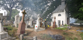 Osoba czyści nagrobek na cmentarzu z kilkoma nagrobkami i małą kapliczką otoczoną drzewami. Teren jest utrzymywany w ramach projektu ochrony polskiego dziedzictwa kulturowego za granicą.