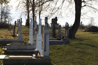 Nagrobki na wiejskim cmentarzu w Bełzie, otoczone trawą i drzewami. Niektóre nagrobki mają krzyże i figury. Cmentarz jest częścią projektu porządkowania i ochrony polskiego dziedzictwa.