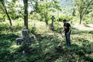 Wolontariusz używa podkaszarki do usuwania zarośniętej trawy wokół starych kamiennych krzyży na cmentarzu w Mołdawii. Teren otaczają drzewa i zieleń.