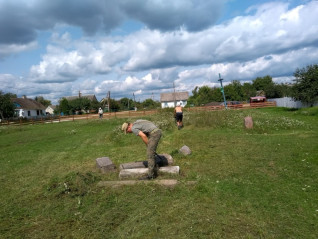 Wolontariusze pracują na cmentarzu na wsi, mężczyzna pochyla się nad grobem. W tle domy, drewniany płot i niebieski krzyż. Niebo jest częściowo zachmurzone.