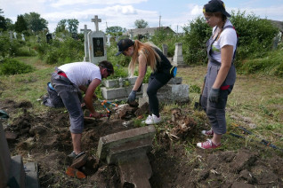 Wolontariusze sprzątają grób na cmentarzu w Bołszowcach na Ukrainie. Dwie osoby klęczą z narzędziami, a trzecia stoi obok. W tle widoczne są nagrobki i zieleń.