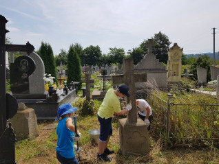 Wolontariusze czyszczą nagrobki na cmentarzu ormiańskim na Ukrainie. Dwie osoby pracują przy kamiennym krzyżu, a inna zajmuje się otoczeniem. W tle widoczne są różne nagrobki i zieleń.