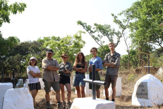 Grupa sześciu młodych wolontariuszy stoi wśród białych grobów z krzyżami na polskim cmentarzu w Zambii. W tle widoczne są drzewa i zieleń.