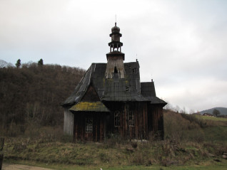Drewniana kaplica z zużytym dachem i wieżą, otoczona wzgórzami i rzadką roślinnością, pod pochmurnym niebem.