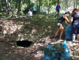 Wolontariusze sprzątają cmentarz w zalesionej okolicy, zbierając liście i śmieci do niebieskich worków. W ziemi częściowo widoczna jest stara konstrukcja z cegieł.