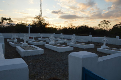 Polski cmentarz z białymi grobami i krzyżami o zachodzie słońca w Afryce, otoczony niskim murem i drzewami w tle.