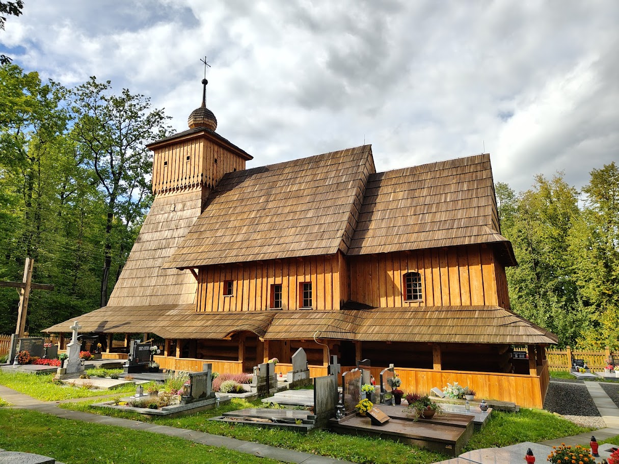 Wooden Corpus Christi Church in Guty with a steep roof and tower, surrounded by a cemetery with gravestones. Trees and a cloudy sky in the background.