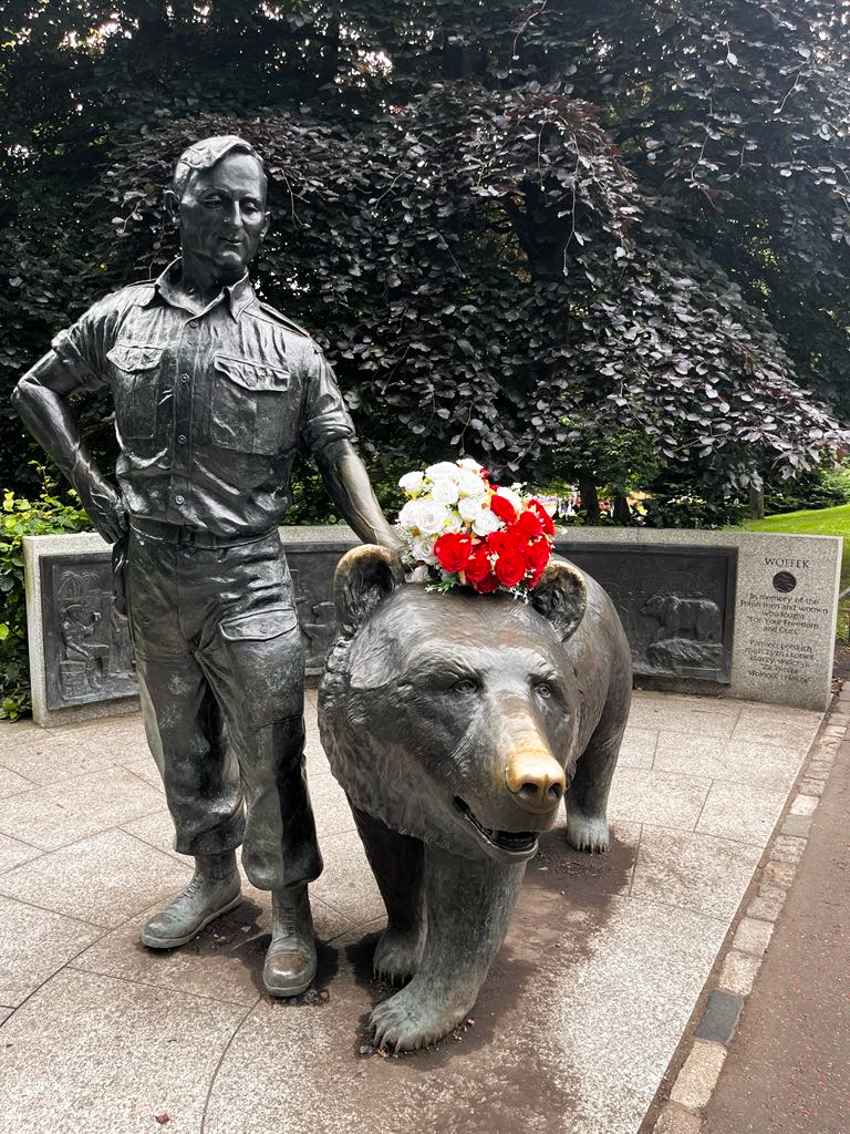 Statue en bronze de l'ours Wojtek et d'un soldat polonais dans les Princes Street Gardens, Édimbourg. Wojtek porte une couronne de fleurs rouges et blanches. Des bas-reliefs illustrant l'histoire de Wojtek sont visibles en arrière-plan.