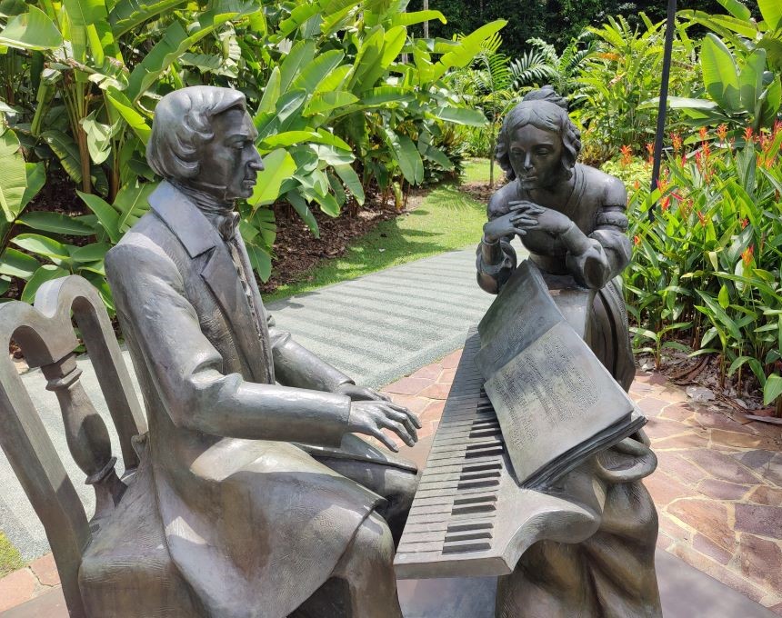 Sculpture en bronze représentant Frédéric Chopin assis à un piano et une femme appuyée contre celui-ci, entourée d'une végétation luxuriante dans les jardins botaniques de Singapour.