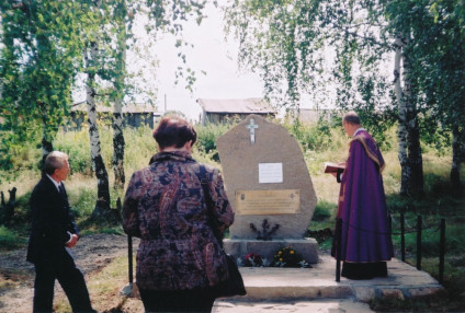 Trzy osoby stoją przy kamieniu pamiątkowym na cmentarzu, otoczonym brzozami. Na kamieniu znajduje się krzyż i tabliczki, jedna osoba w fioletowej szacie trzyma książkę.