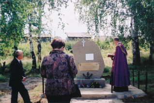 Trzy osoby stoją przy kamieniu pamiątkowym na cmentarzu, otoczonym brzozami. Na kamieniu znajduje się krzyż i tabliczki, jedna osoba w fioletowej szacie trzyma książkę.