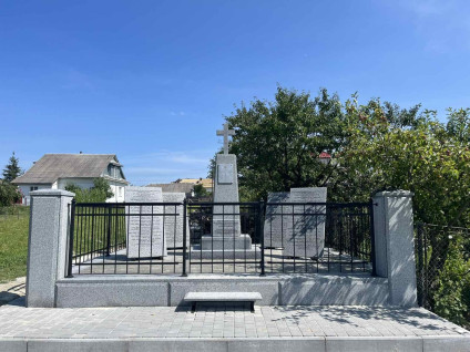 Grób zbiorowy legionistów polskich w Mołotkowie, otoczony metalowym ogrodzeniem. W centrum stoi kamienny obelisk z krzyżem, otoczony tablicami z inskrypcjami. W tle widoczne drzewa i domy.