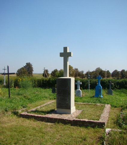 Betonowy krzyż na prostym cokole oznacza grób 18 polskich żołnierzy poległych we wrześniu 1939 roku, położony na trawiastym terenie z innymi grobami w tle.