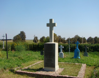 Betonowy krzyż na prostym cokole oznacza grób 18 polskich żołnierzy poległych we wrześniu 1939 roku, położony na trawiastym terenie z innymi grobami w tle.