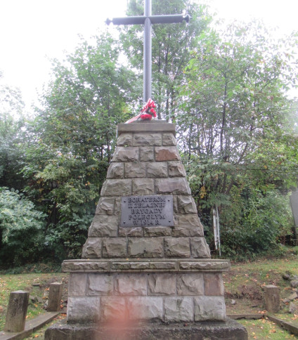 Kamienny obelisk z żeliwnym krzyżem pomalowanym na srebrno, poświęcony bohaterom II Żelaznej Brygady poległym w 1914 roku. Otoczony łańcuchami i betonowymi słupkami, na tle drzew.