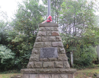 Kamienny obelisk z żeliwnym krzyżem pomalowanym na srebrno, poświęcony bohaterom II Żelaznej Brygady poległym w 1914 roku. Otoczony łańcuchami i betonowymi słupkami, na tle drzew.