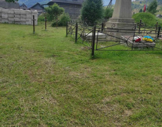 Pomnik II Brygady Legionów Polskich w Rafajłowej. Kamienny obelisk z krzyżem na szczycie, otoczony metalowym ogrodzeniem. W tle drewniany dom i wzgórza.