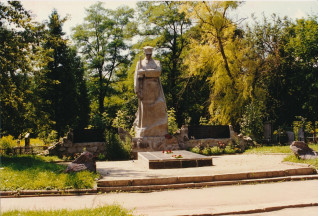 Kamienna figura żołnierza w centrum kwatery wojennej w Sumach, otoczona drzewami. Dwie tablice w językach polskim i ukraińskim znajdują się na niskim murku za figurą. U podstawy leżą kwiaty.
