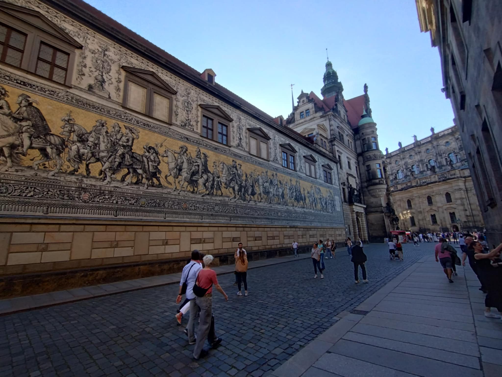 Orszak książęcy na ścianie Langer Gang w Dreźnie, wykonany z porcelanowych płytek, przedstawiający władców Saksonii, w tym królów Polski. Ludzie spacerują wzdłuż brukowanej ulicy obok malowidła.